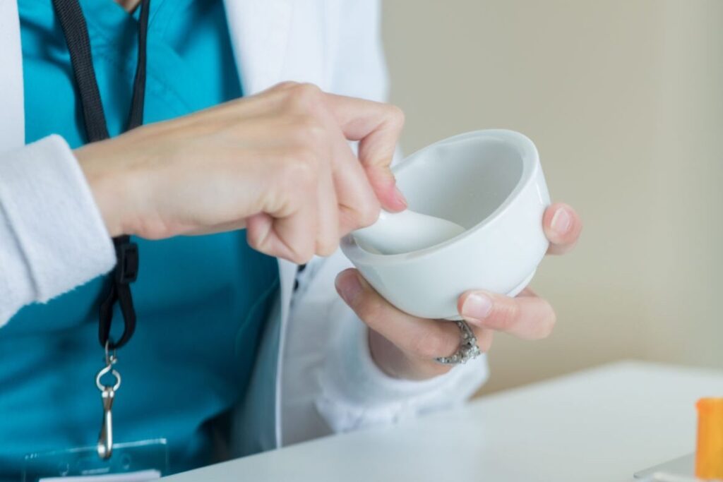 This closeup focuses on a mortar and pestle as an unrecognizable, seen from the torso, pharmacist sits at her desk and compounds medication to fill a prescription. There is a laptop, prescription bottle and pill counter on the desk.